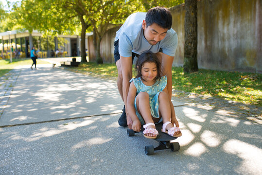 Portrait of Asian dad and girl resting with skateboard in park. Bearded man in casual clothes bending over daughter pushing skateboard while kid sitting on it. Healthy rest and happy childhood concept