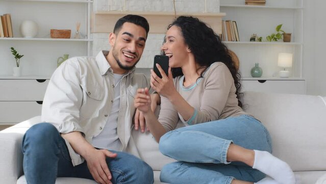 Multiracial family Indian Hispanic man and ethnic Latina Caucasian woman couple sitting on sofa at home trying photo mask in mobile app watching funny video in phone fun with smartphone talking laugh