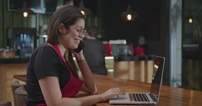 Female hispanic barista employee working in front of laptop computer taking online delivery. Young woman using technology speaking with client and multi tasking