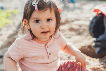 Close-up portrait of a young baby girl sitting in the sand on the backyard, looking at camera and smiling. Outdoor lifestyle. Natural beauty. Smile emotions