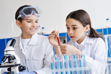 amazed girl holding test tube with red liquid near smiling boy and microscope in lab.