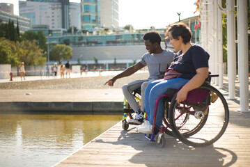 Happy man and woman in wheelchairs spending time near water. African American man and Caucasian woman in casual clothes, looking at water, discussing something. Love, affection, happiness concept