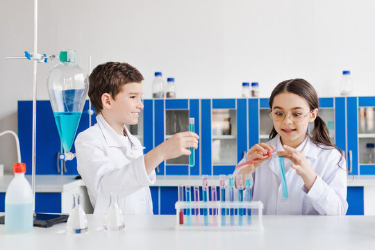 Happy Preteen Kids In White Coats Looking At Camera Near Microscope And Test Tubes With Flasks.