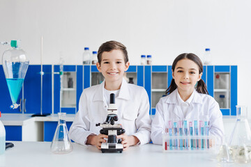 happy preteen kids in white coats looking at camera near microscope and test tubes with flasks.