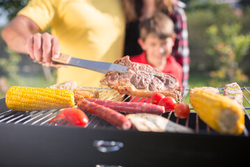Happy father grilling meat and vegetables with children. Dark-haired man in yellow T-shirt putting meat on BBQ grid. Son and daughter standing next to him. BBQ, cooking, food, family concept