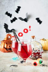 Halloween bloody cocktails. Two bloody cocktails glass goblets for Halloween and bottle form skull on light backgrounds. Decor concept for Halloween party with candy eyes, bats, spiders and pumpkins.