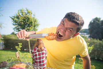Funny man at BBQ family party. Dark-haired man in yellow T-shirt eating freshly roasted meat and...