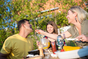 Joyful family eating BBQ at backyard. Mid adult parents and daughter sitting around table, eating freshly cooked food. Wife feeding husband. BBQ, cooking, food, family concept