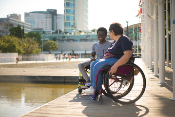 Laughing man and woman in wheelchairs spending time near water. African American man and Caucasian woman in casual clothes, looking at water, joking. Love, affection, happiness concept