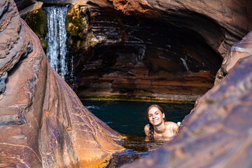 A girl in a white bikini relaxes in a rock pool with a waterfall cascading over her head; swimming in natural pools in karijini national park, western australia
