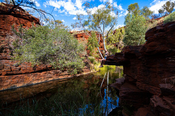 Fototapeta na wymiar A jungle girl in a bikini sits on rocks by a stream in a canyon in karijini national park, western australia; an oasis in the desert in the australian outback, terra rosa, australia