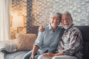 Cute and beautiful couple of old people smiling and looking at the camera having fun at home together. Portrait of seniors wearing eyeglasses sitting on sofa enjoying and relaxing..