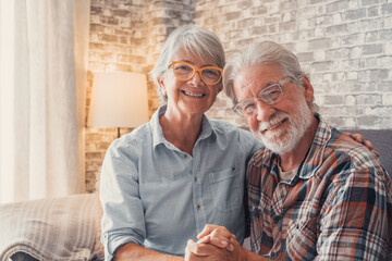 Cute and beautiful couple of old people smiling and looking at the camera having fun at home together. Portrait of seniors wearing eyeglasses sitting on sofa enjoying and relaxing..