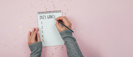 Woman's hand writing 2023 goals in note pad on pink background. Top view. Concept of New Year's plans, goals and actions