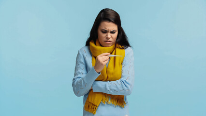 sad and sick woman in scarf holding electronic thermometer isolated on blue