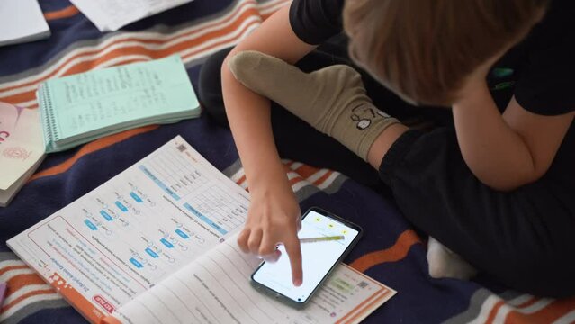 Close up preteen child doing homework exercise sit on bed at home. Using smartphone searching information. Mess in children room. School classes for secondary. Remote education. E-learning. Top view