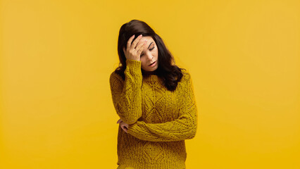 upset and brunette woman in sweater touching forehead isolated on yellow