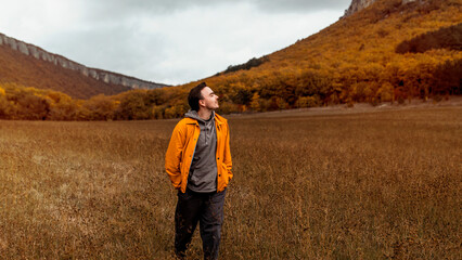 Fototapeta na wymiar Young smiling millennial man in a yellow jacket walks near the picturesque mountains and autumn forest in October