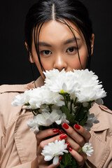 Stylish asian woman in trench coat covering face with flowers isolated on black