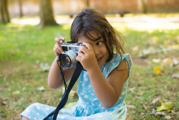 Close-up of little Asian girl taking photos on digital camera. Happy dark-haired child sitting on grassy ground holding camera and looking into it to take pictures. Leisure and happy childhood concept