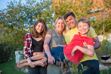 Fototapeta premium Portrait of funny family at backyard. Mid adult couple and children. Holding smiling boy together, making faces. BBQ, love, food, family concept