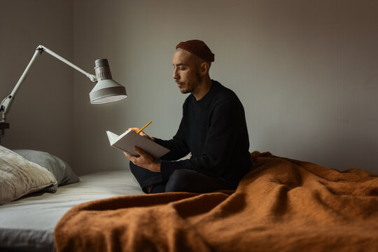 A caucasian man wearing an orange beanie hat sitting in a bed writing in a book with a bed lamp on.