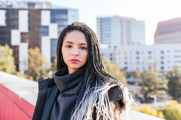 Serious Hispanic female in warm outerwear with dreadlocks looking at camera while standing near border of residential building in city