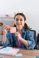 cheerful girl showing empty sticky note and looking at camera at home.