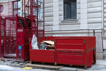 Red metal dumpster container full of construction garbage near freight construction elevator, house renovation.