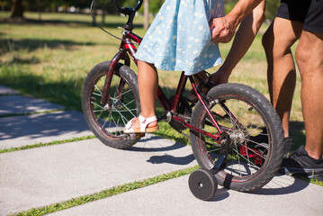 Close-up of little girls legs riding small bike in summer. Girl in blue dress sitting on bicycle and man holding it walking by daughters side. Active rest and healthy growing-up concept