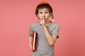 a boy in a construction helmet and a gray t-shirt put his finger to his mouth, holds a book in one hand and stands on a pink background