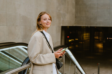 Young white woman using cellphone while standing by escalator outdoors