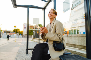 Young woman smiling and using cellphone while sitting at bus station