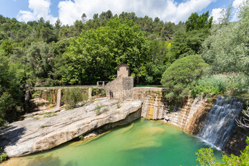 Fototapeta na wymiar Half-height view of the old power plant by a waterfall in a natural park that is transformed into a pond. The river falls like a waterfall into a green pond from a stone wall full of vegetation 