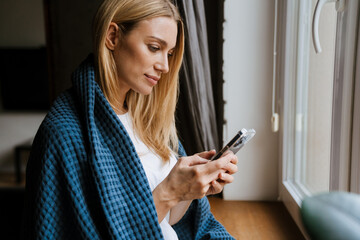 Blond white woman using cellphone while standing by window at home