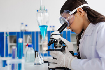 side view of preteen girl in goggles looking into microscope in chemical laboratory.