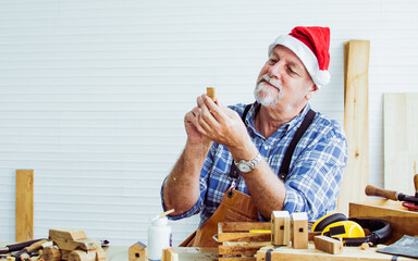 Portrait senior old aging kind Caucasian man or carpenter wearing santa hat, making DIY wooden furniture, holding eyeglasses, smiling with happiness, sitting alone at home. Retirement, Hobby Concept.