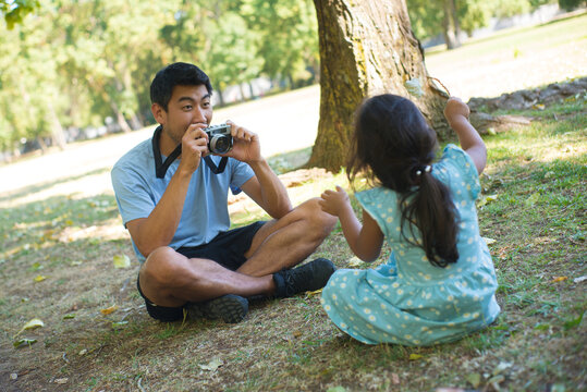 Asian father getting ready to take photo of his kid on field. Happy dad holding camera and his little daughter sitting on green field and posing for camera. Leisure and happy childhood concept