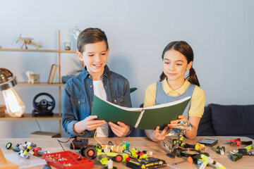 Smiling kids holding notebook near robotic model and tools at home.