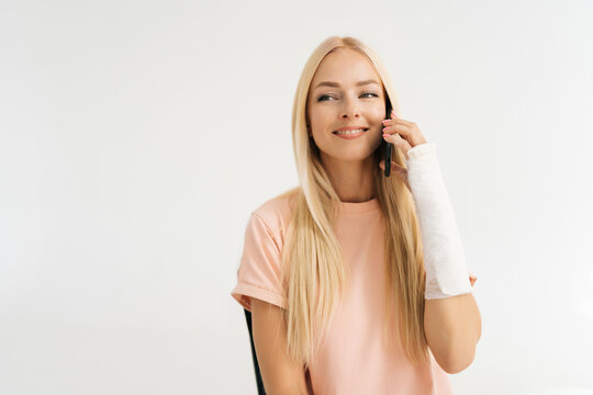 Studio portrait of cheerful injured blonde young woman with broken arm wrapped in plaster bandage talking smartphone on white isolated background, looking away. Concept of insurance and healthcare.