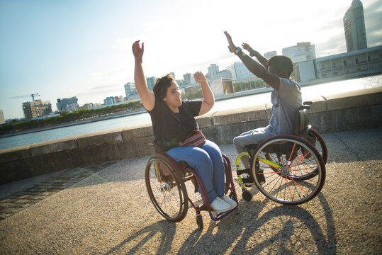 Jolly biracial couple having walk on summer day. African American man and Caucasian woman in wheelchairs on embankment, fooling around. Love, relationship, happiness concept - Powered by Adobe