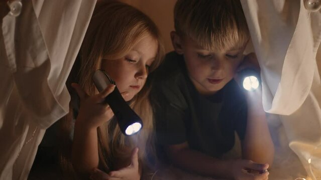 Happy family. Two children girl and boy are reading book in tent. Flashlight in hands of brother and sister. Happy family with book in tent. Children holds. reading book in selfmade house at bedroom