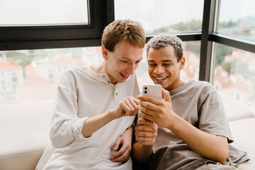 Happy gay couple using cellphone together while resting on sofa at home