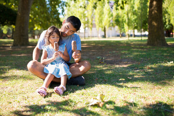 Portrait of Asian father and girl having good day in park. Happy man in casual clothes and his daughter sitting on grass under trees and eating ice cream. Fatherhood and leisure with kids concept