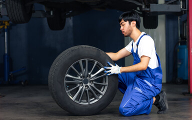 Naklejka na ściany i meble Asian handsome male mechanic wearing uniform, changing rubber tyre or tire wheel, working, reparing in garage at car or automobile maintenance service center or shop with copy space. Industry Concept.