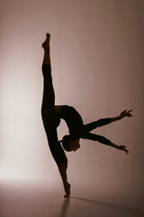 the silhouette of a rhythmic gymnastics girl in a bodysuit shows an exercise in vertical splits