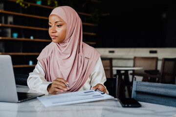 Young muslim woman wearing headscarf smiling while working in office - obrazy, fototapety, plakaty