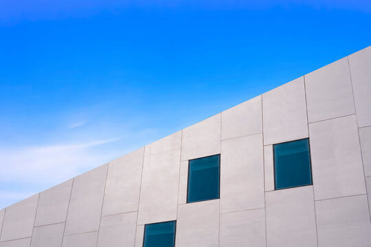 Low Angle And Side View Of Glass Windows On Modern White Concrete Building Wall Against Blue Sky, Street Style And Minimal Exterior Architecture Background