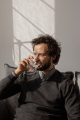 The man drinks water sitting on the sofa. Portrait of a young curly man. Cozy apartment and sunlight