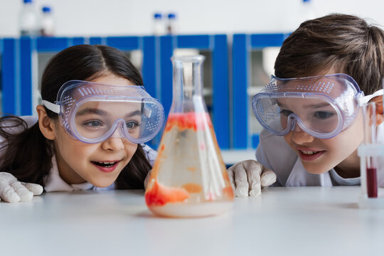 amazed kids in goggles looking at blurred flask with liquid while doing chemical experiment in lab.